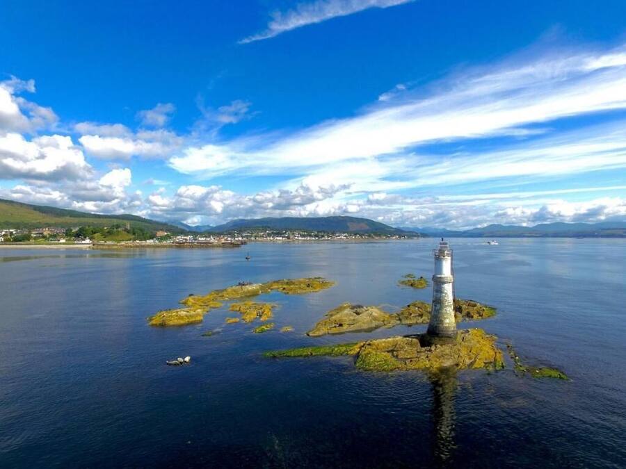 A lighthouse on a rocky island surrounded by calm water, with a distant shoreline and mountains under a partly cloudy sky.