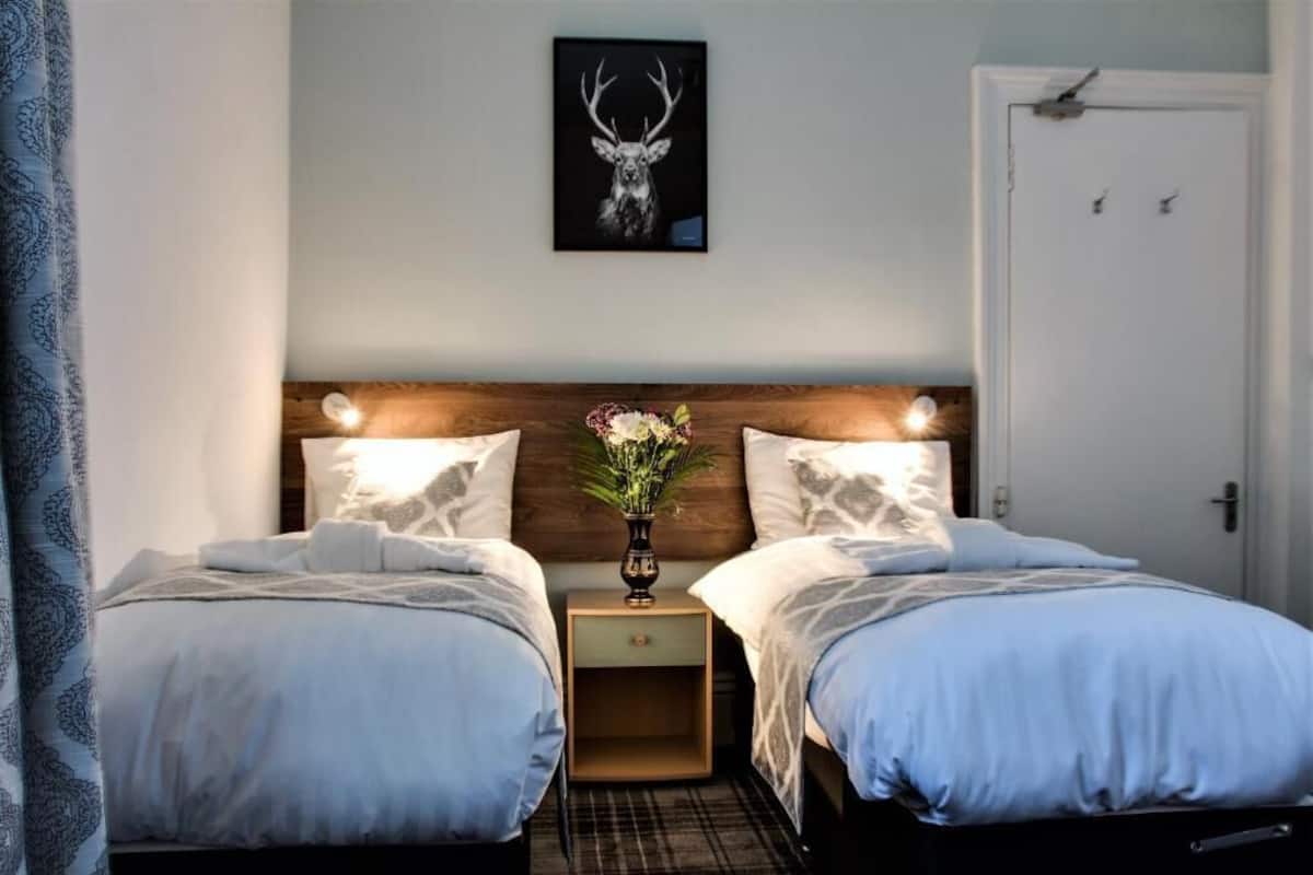 A hotel room with two beds, a wooden headboard, a framed deer antler artwork, and a nightstand with a vase of flowers.