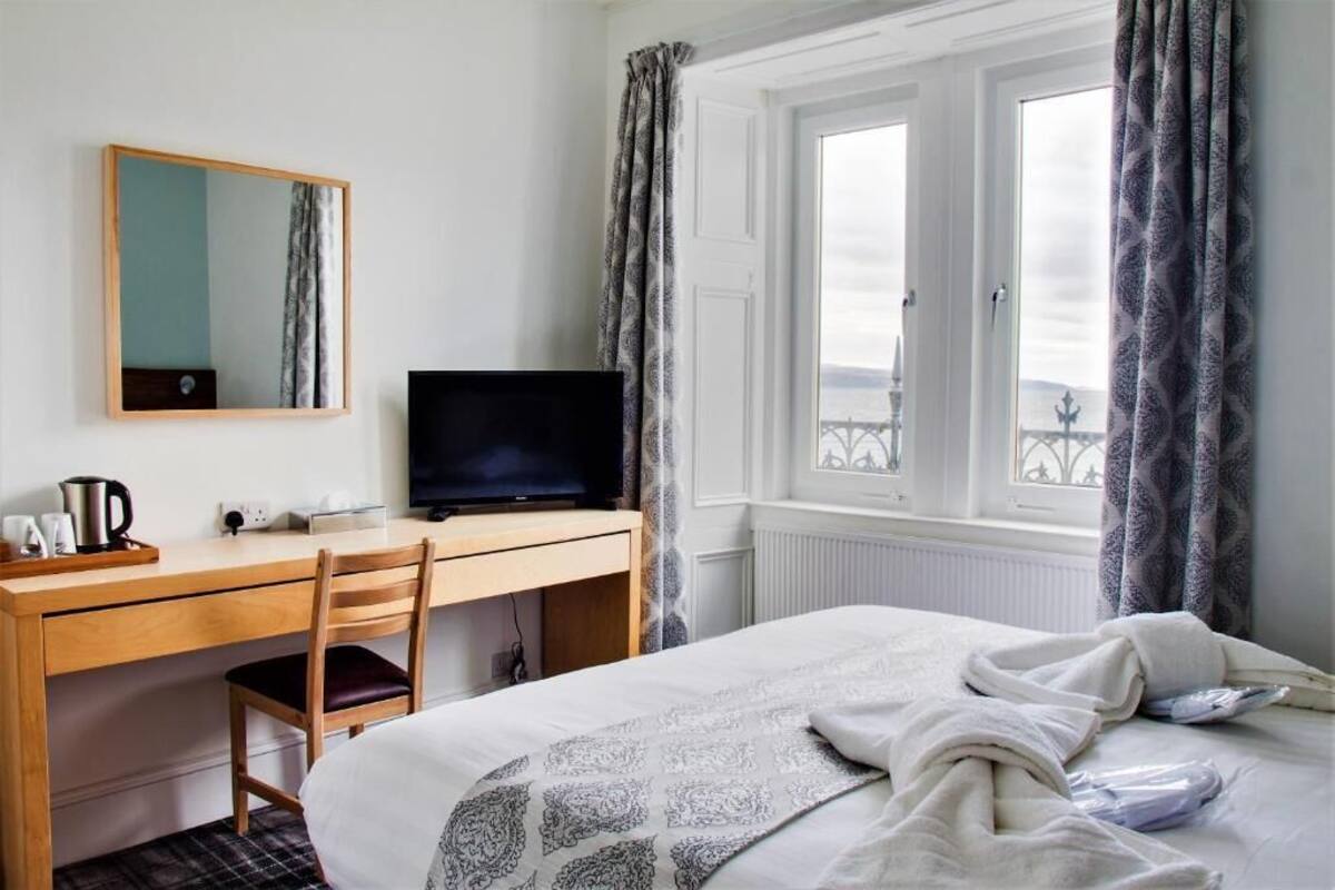 A hotel room with a bed, a desk with a computer, a chair, a window with curtains, and a view of a bridge.