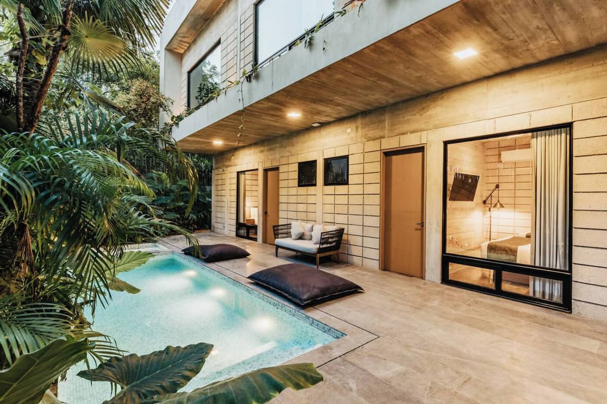 A modern outdoor pool area with a lounge chair and cushions, surrounded by lush tropical plants.