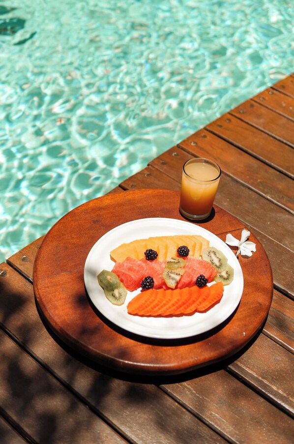 A wooden table with a plate of fruit and a glass of juice by a pool.