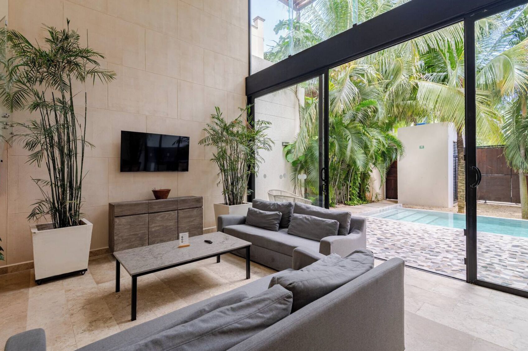A modern living room with a grey sofa, a coffee table, and a TV on the wall. There are large windows with a view of a pool and palm trees.