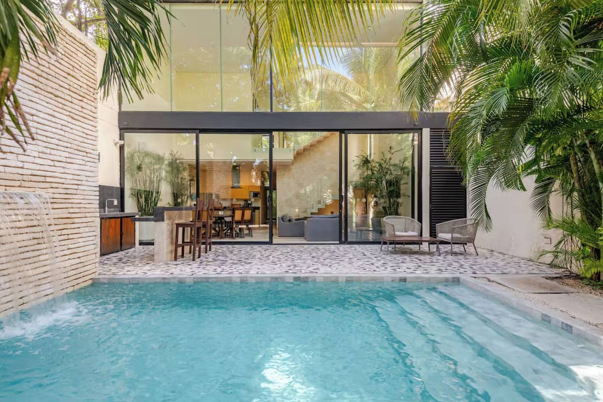 A modern outdoor pool area with a glass-enclosed dining space, surrounded by lush greenery.