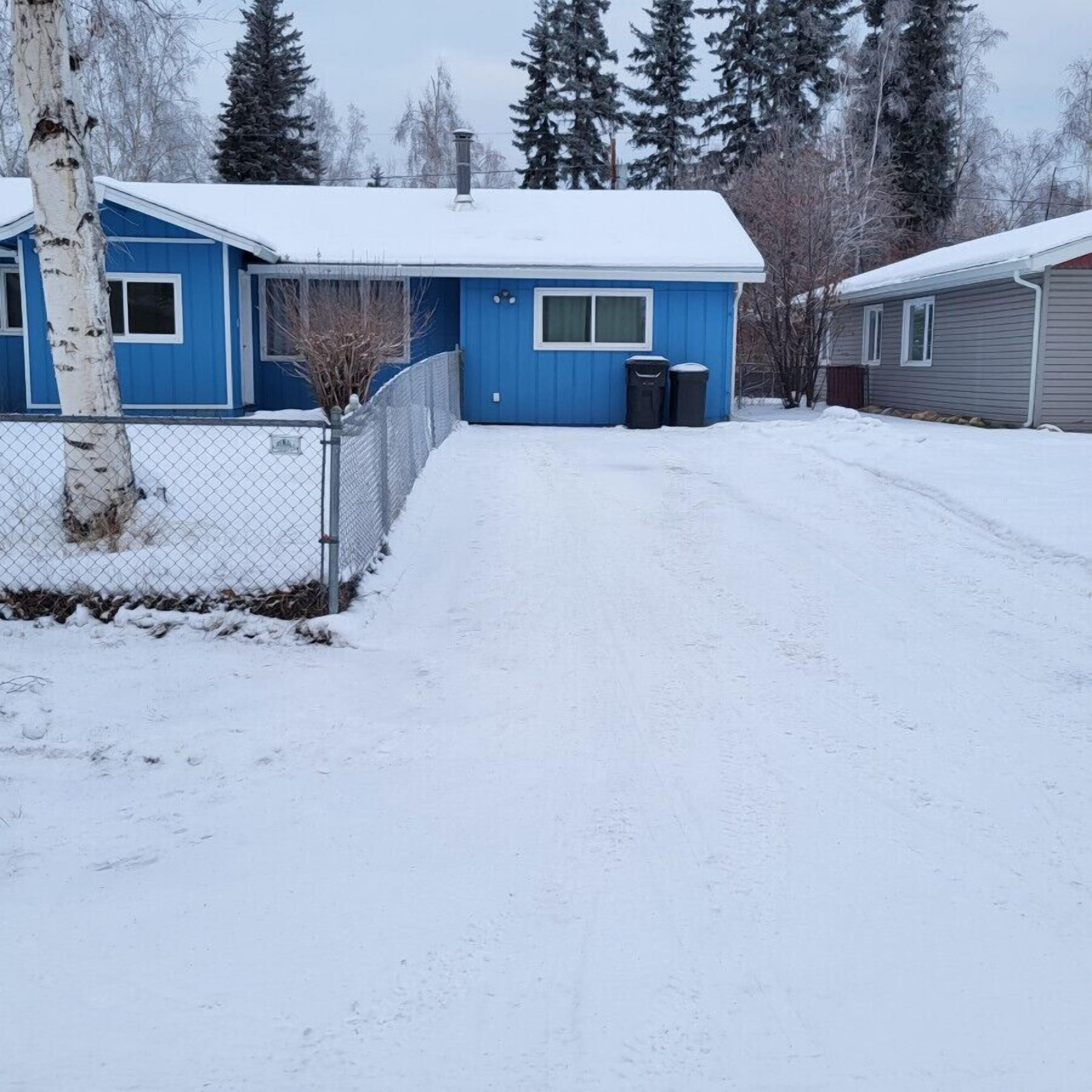 Lumipeitteinen asuinalue, jossa on sininen talo, aidattu piha ja taustalla harmaa rakennus.