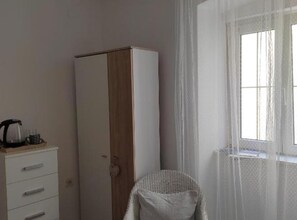 Et værelse med et skab, en stol, et vindue med gardiner og en kaffemaskine på et bord.