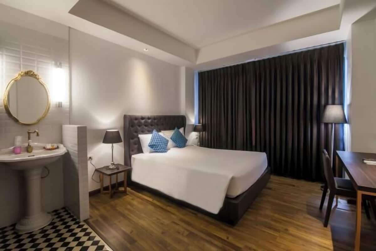 A hotel room with a large bed, a bedside table, a lamp, a mirror, a sink, and a checkered floor.
