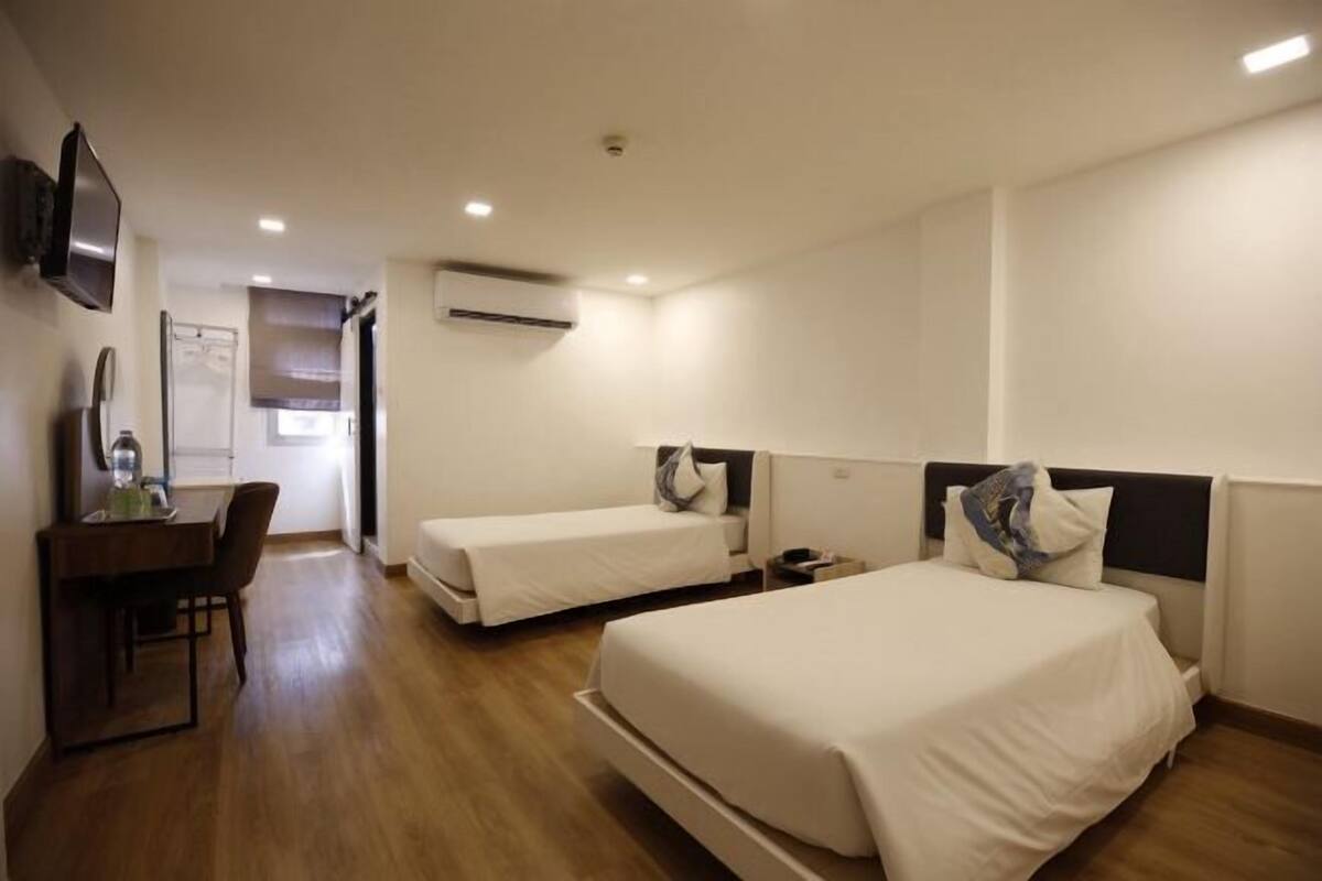 A hotel room with two single beds, a desk with a chair, a flat-screen TV, and an air conditioning unit.