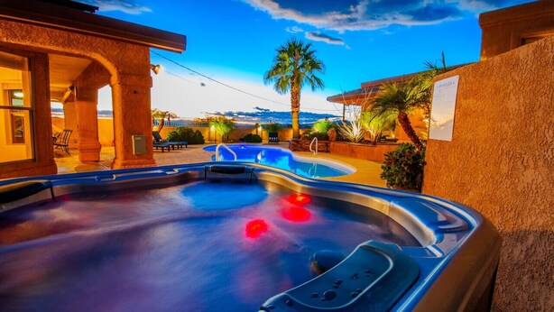 A hot tub area with a pool, palm trees, and a building with a stucco exterior. - Havasu Whaler -  Pool, Spa, Game Room, Casita (Lake Havasu City)