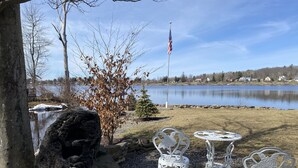 A lakeside patio with two white metal chairs and a small table, surrounded by rocks and a tree with brown leaves. - Cozy cottage-breathtaking sunsets. Lake front. Close to casino & racetrack. (Delanson)