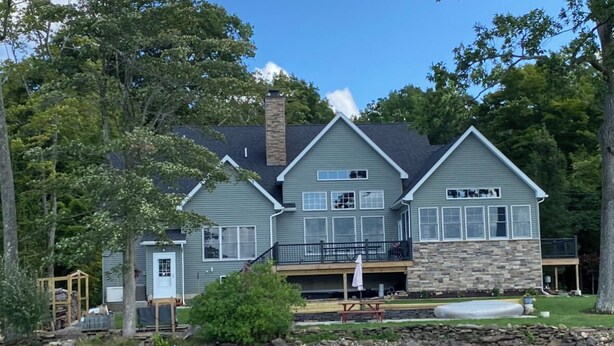 A large, two-story house with a stone foundation and a balcony, situated by a body of water. - Cozy cottage-breathtaking sunsets. Lake front. Close to casino & racetrack. (Delanson)