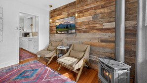 A cozy room with a wooden wall, a fireplace, two chairs, a small table, and a kitchen area in the background.