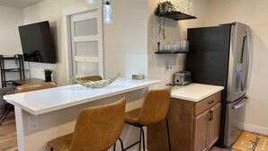 A modern kitchen with a white countertop, two brown bar stools, a stainless steel refrigerator, and a shelf with glasses and plants.