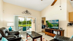 A spacious living room with a large TV, leather sofas, a coffee table, and a view of a pool through glass doors.