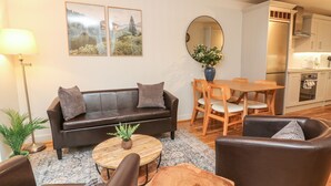 Un salon moderne avec un canapé, une table basse et des fauteuils. On y trouve également un coin repas avec une table et des chaises. Une cuisine équipée, avec des placards et des appareils électroménagers, est visible au fond.