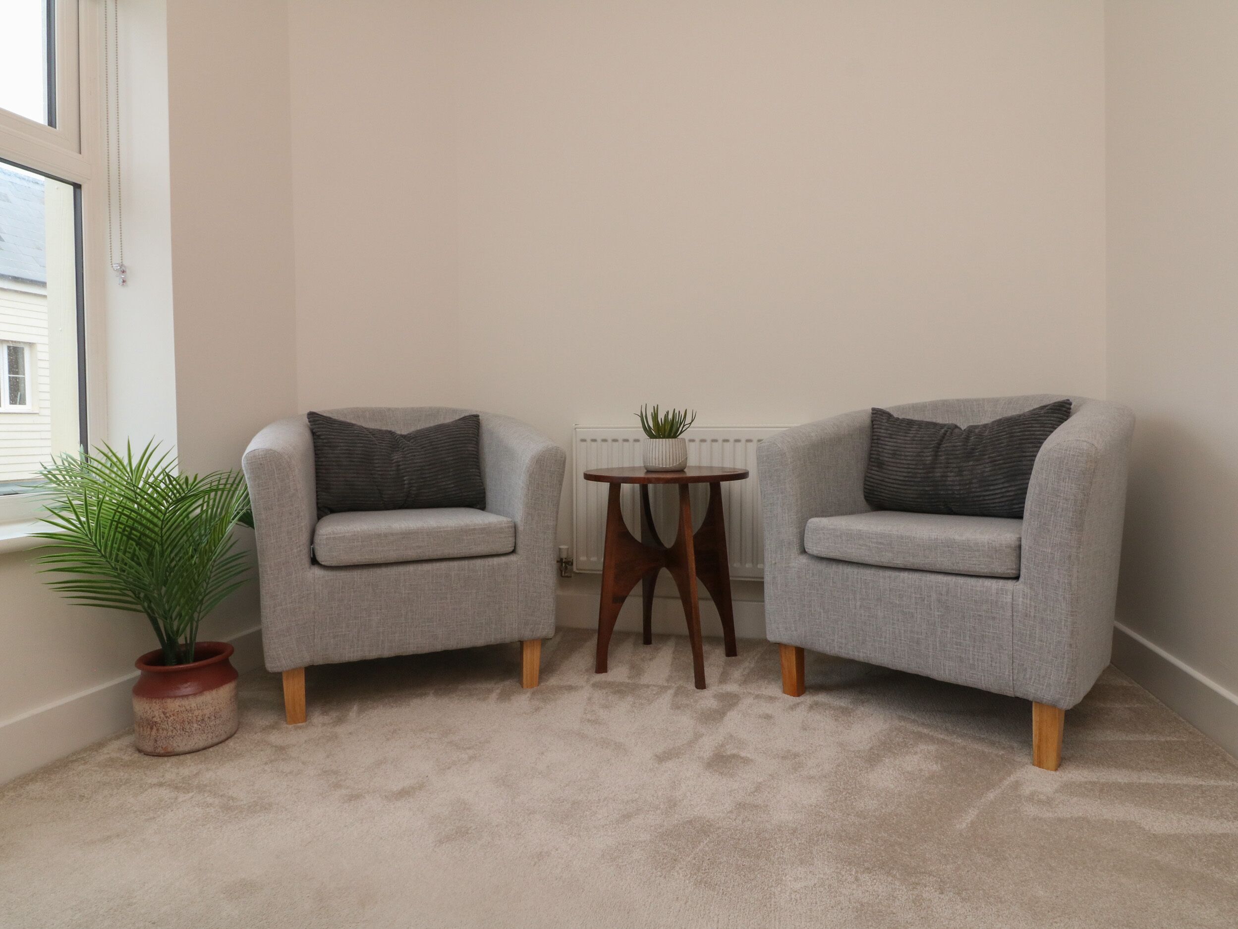 A room with two grey armchairs, a small wooden table, a potted plant, and a radiator.
