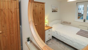 Une chambre à coucher comprenant un lit, une table de chevet, un miroir, une armoire en bois, une petite plante et une fenêtre donnant sur des immeubles.