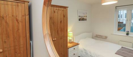 A bedroom with a bed, a nightstand, a mirror, a wooden wardrobe, a small plant, and a window with a view of buildings.