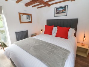 A neatly made bed with a grey blanket, red pillows, and a white duvet. There is a framed picture on the wall above the bed, a bedside table with a lamp, and a radiator on the wall. - Tor Barn (Matlock)