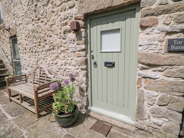 A stone building with a green door, a bench, and a potted plant with purple flowers. - Tor Barn (Matlock)
