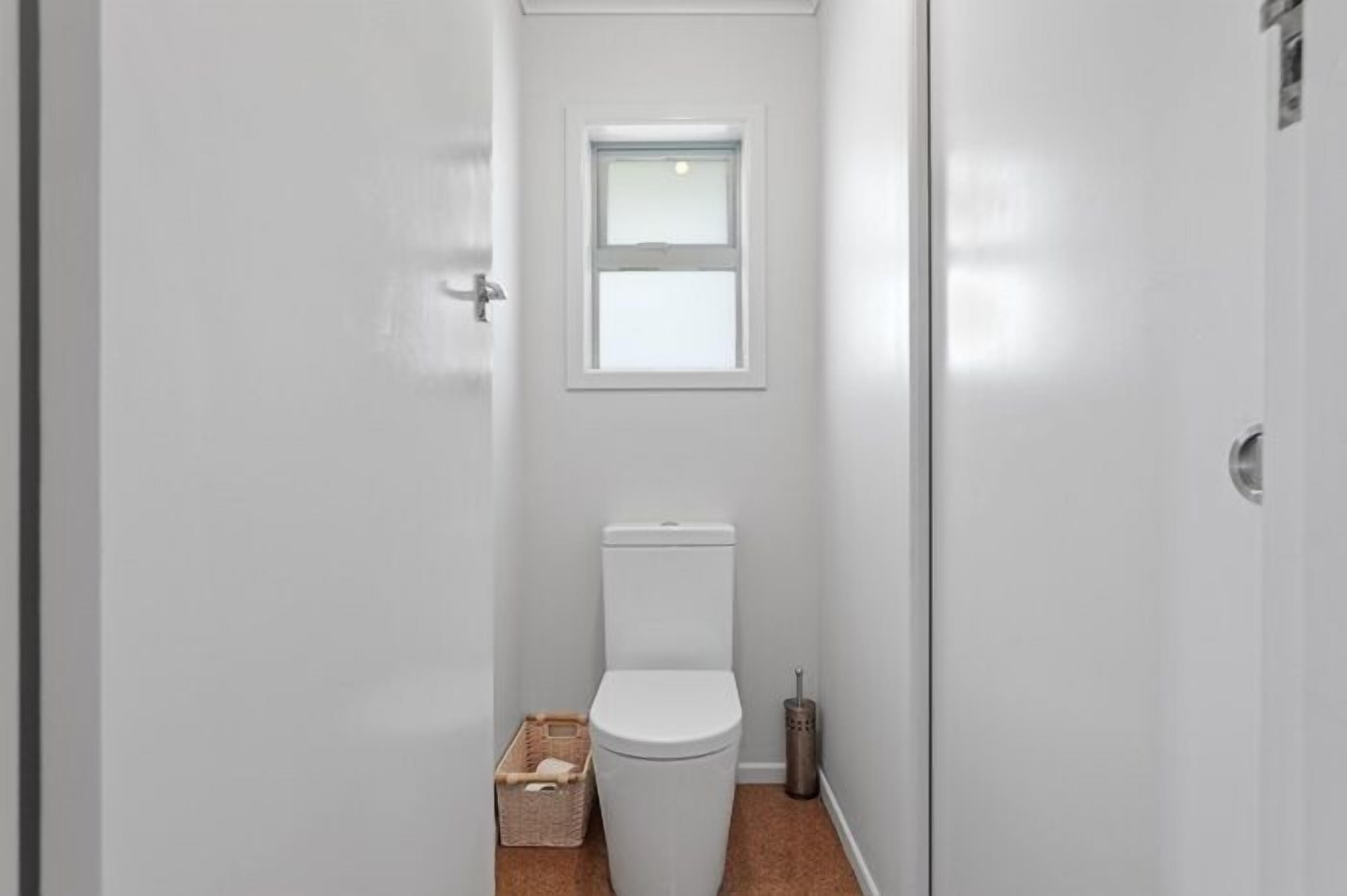 A narrow bathroom with a white toilet, a small window, and a broom.