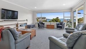 A modern living room with grey leather sofas, a flat-screen TV, and a large window offering a view of the sea and buildings. - Home Away From Home - Taupo (Taupo)