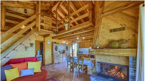Ein gemütliches Blockhaus-Interieur mit Kamin, Holzbalken, einer Küchenzeile und einer Couch mit farbenfrohen Kissen.