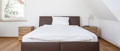 Un dormitorio moderno con una cama grande, cajones de madera, una mesita de noche y una lámpara.
