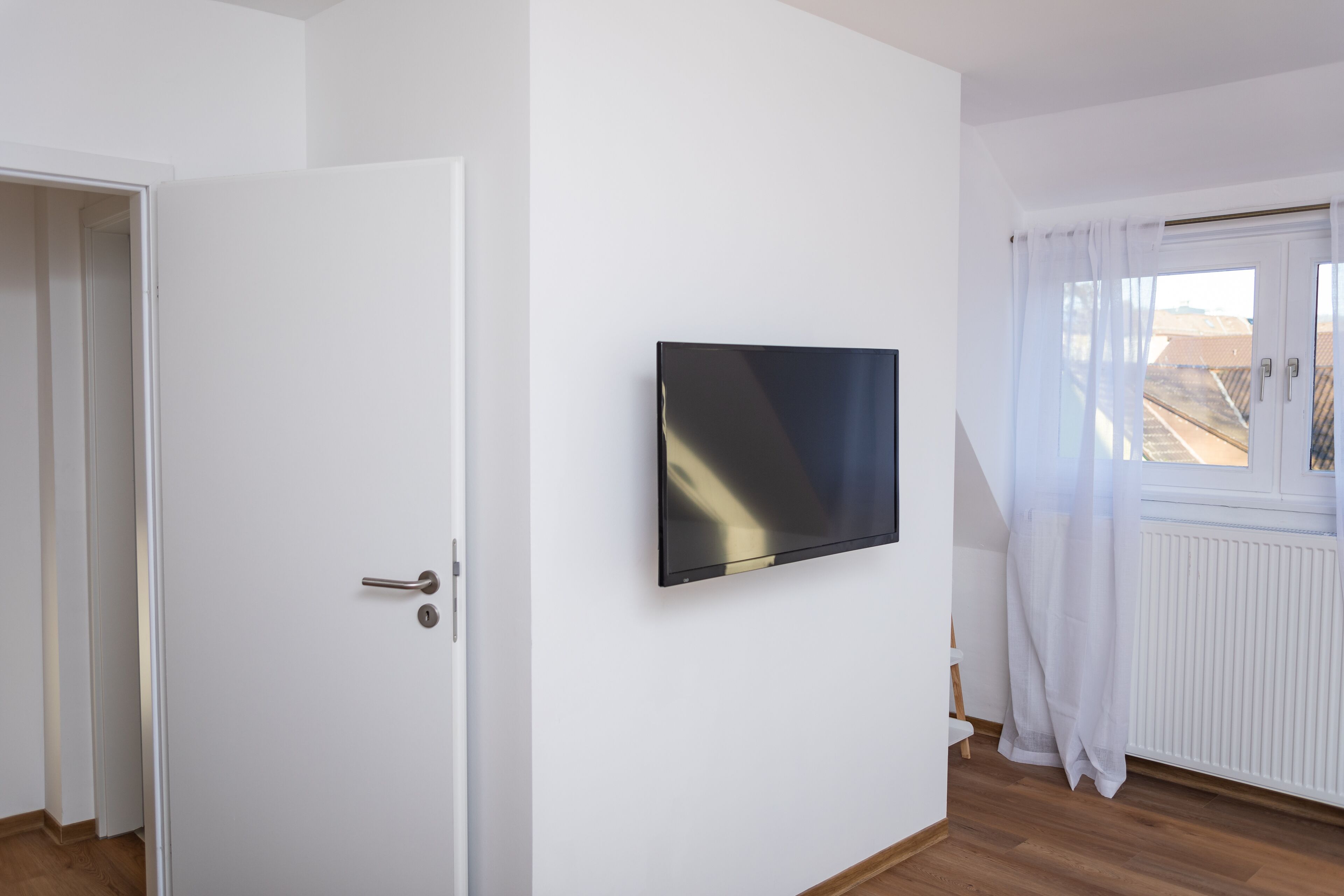 A room with a white door, a mounted flat-screen TV, a window with white curtains, and a radiator.