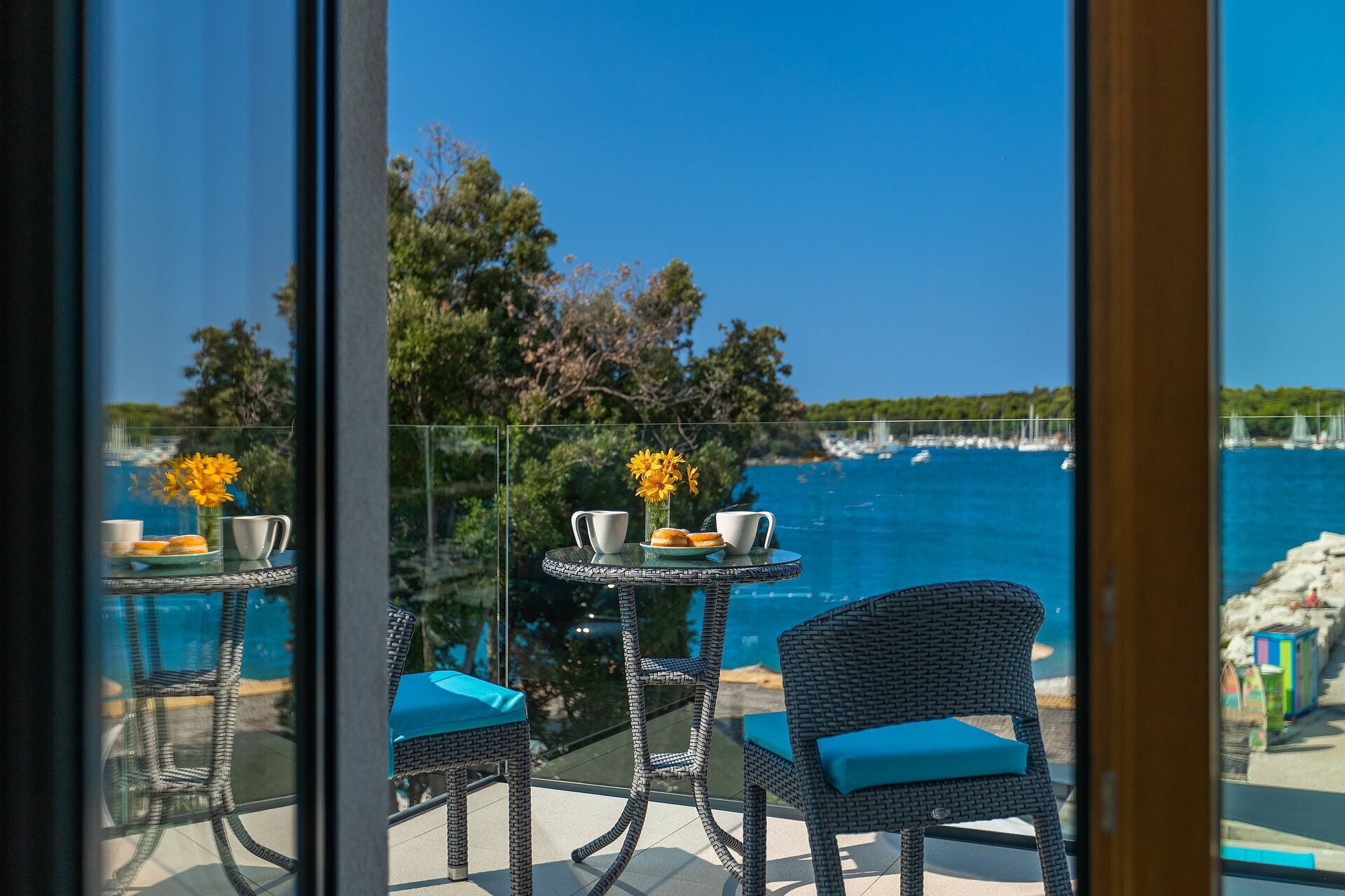Un balcón con sillones y mesitas de mimbre, con vista a un mar azul claro y algunas embarcaciones.