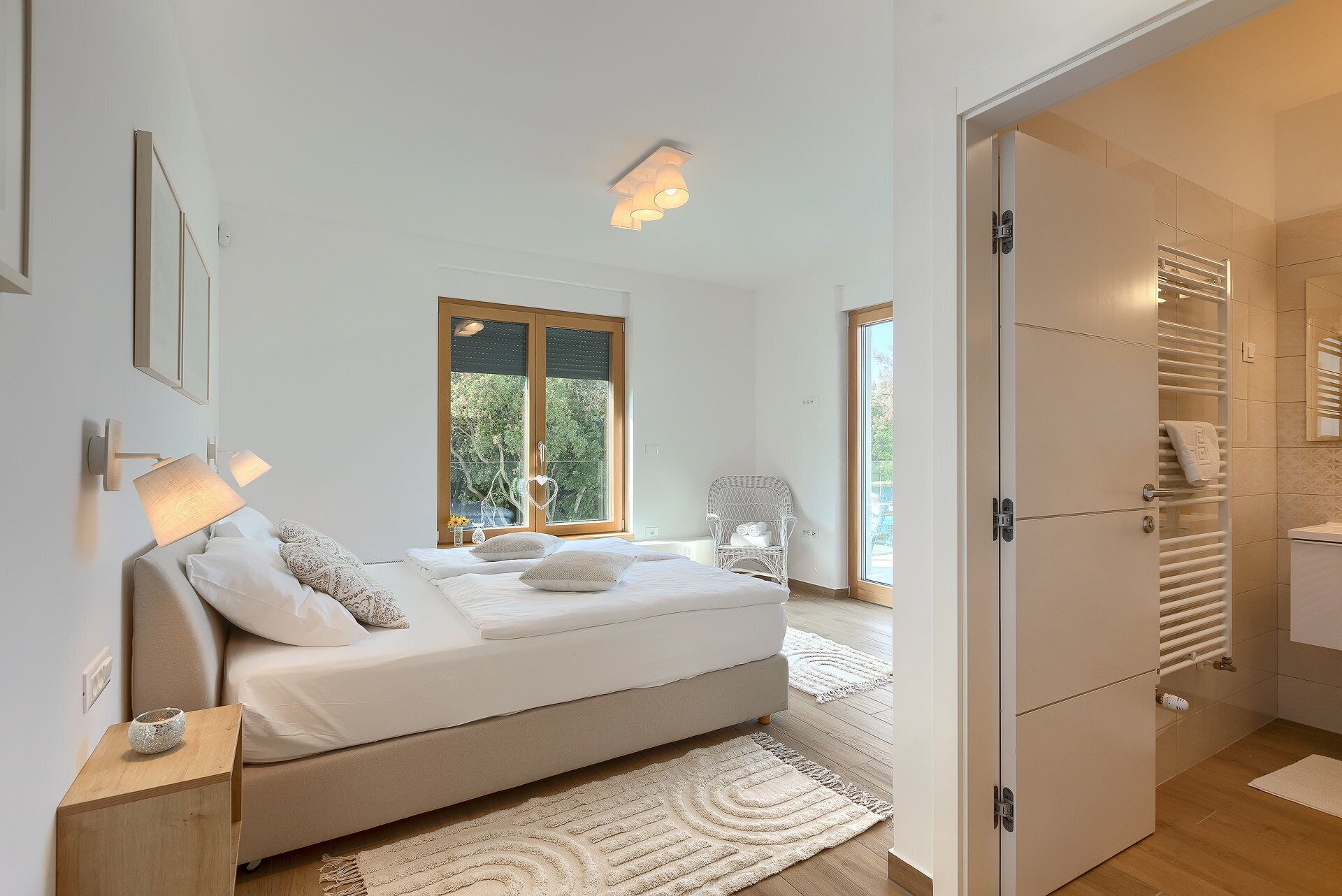 Un dormitorio moderno con cama, lámparas de noche, una silla y vistas al exterior con vegetación.