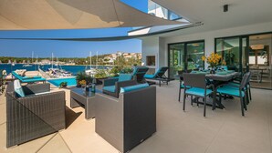 A sunlit outdoor patio with wicker furniture, a dining table set for two, and a view of the marina and boats. - Beachfront Villa Naviblue with Pool (Pula)