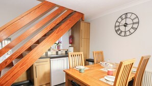 A modern kitchen with wooden cabinets, a built-in dishwasher, and a dining area with a wooden table and chairs. A large wall clock is visible above the dining table. - Stoneleigh Cottage (Richmond)