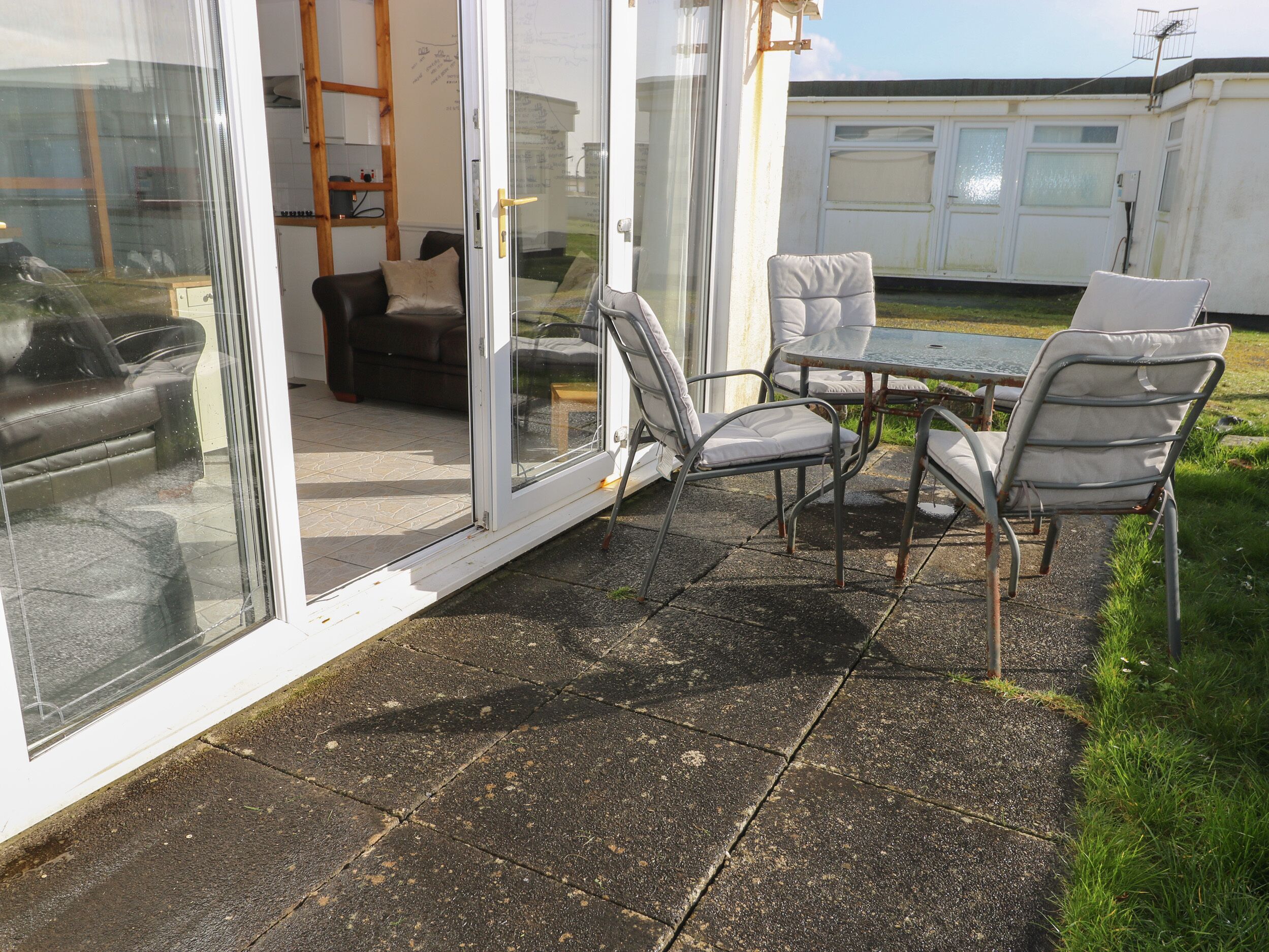A patio area with a glass door leading to a living room, a table with chairs, and a view of a caravan in the background.