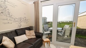 A living room with a black leather sofa, two cushions, a small wooden table, and a glass door leading to an outdoor patio with chairs and a table. - Shipwreck Chalet (Kidwelly)