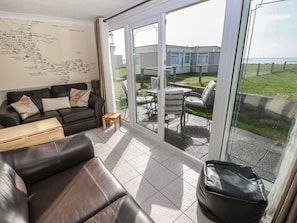A modern living room with a leather sofa, a small wooden table, and a glass door leading to an outdoor patio with a view of the sea.