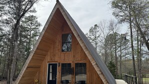 Une cabane triangulaire en bois, dotée d’une porte, de deux fenêtres et d’un petit porche avec deux chaises.