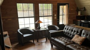 L’intérieur chaleureux d’un chalet avec un plancher en bois, un canapé en cuir, deux fauteuils, une petite table et un coin cuisine équipé d’une cuisinière.