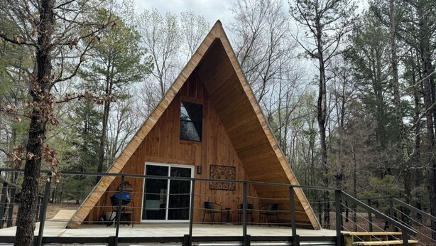 Une cabane triangulaire en bois, dotée d’une porte en verre, entourée d’arbres.