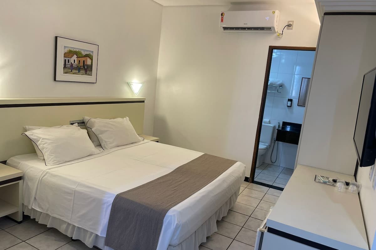 Une chambre d’hôtel avec un lit, une table de chevet et un petit bureau avec un miroir. Il y a un tableau accroché au mur et un appareil de climatisation au-dessus du lit.