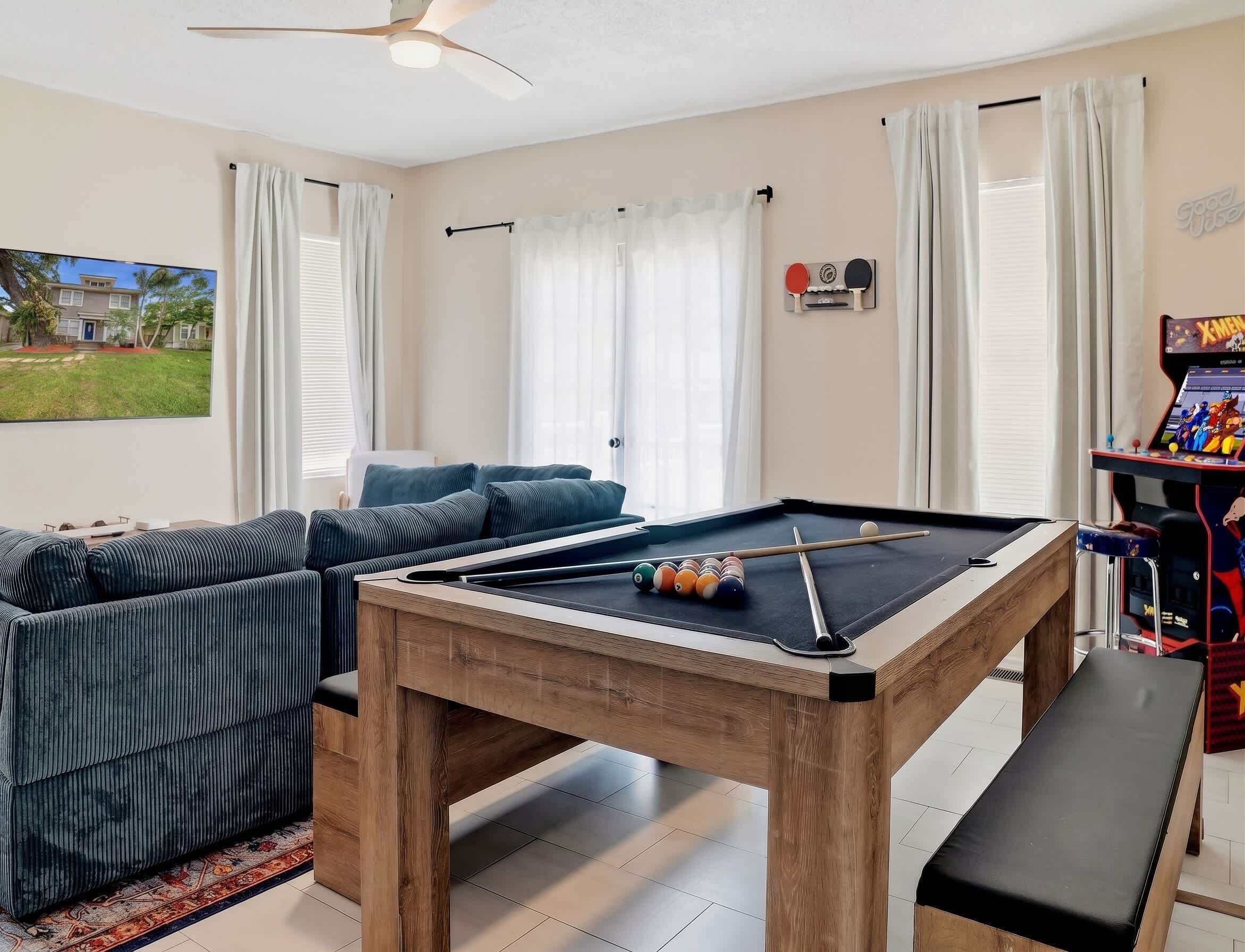 A pool table with balls, a sofa, a bench, a ceiling fan, and a view of a house through a window.