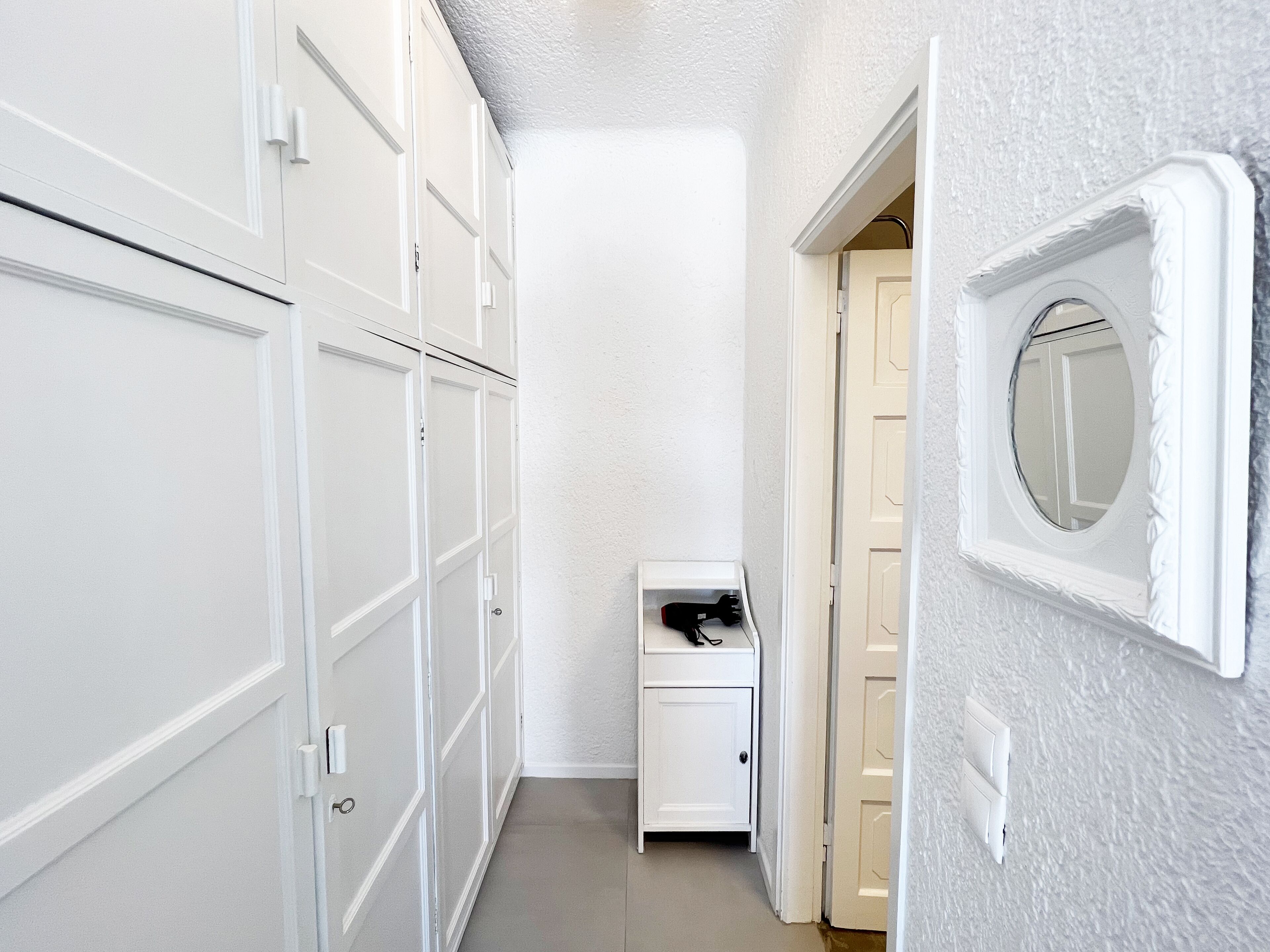 A narrow hallway with white lockers on one side and a small white cabinet with a mirror on the other.