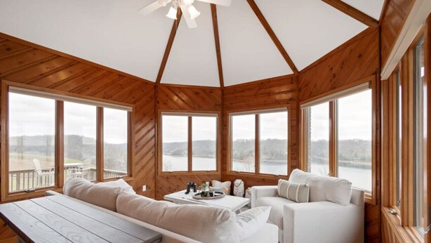 Uma cabana com interior de madeira, janelas amplas, um ventilador de teto e uma área de estar com mesa.