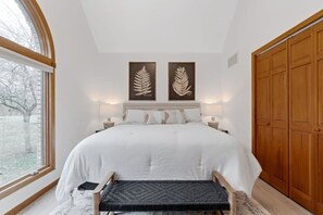 A bedroom with a large bed, two wall-mounted lamps, a wooden wardrobe, and a window with a view of trees.