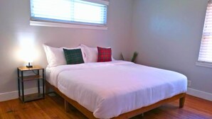 Un dormitorio con cama de madera, ropa de cama blanca, una mesita de noche con lámpara y una ventana con persianas.