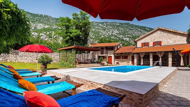 A pool area with lounge chairs and umbrellas, a stone building with a tiled roof, and a mountainous backdrop.