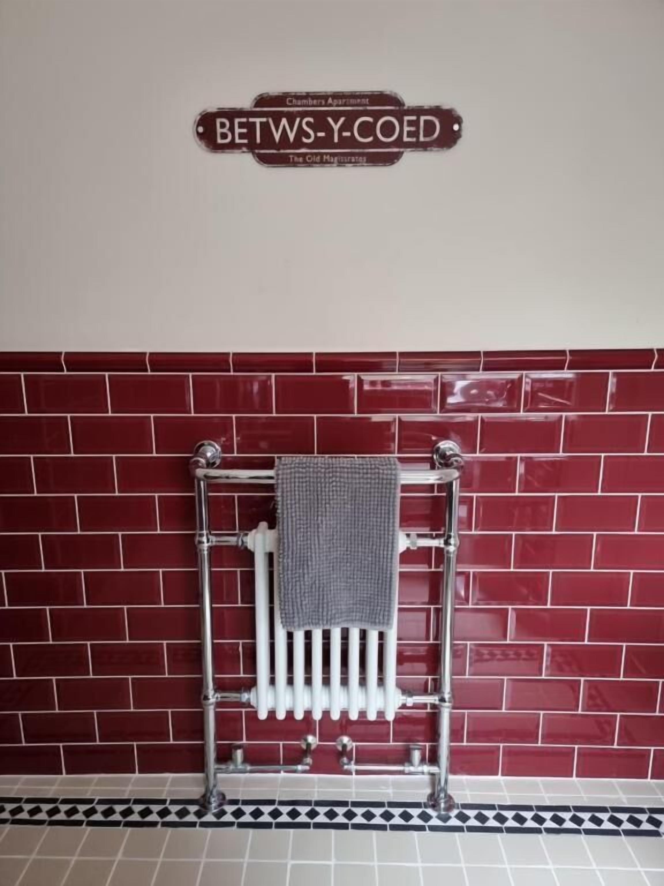 A bathroom with a red-tiled wall, a towel rack with a towel, and a sign above indicating 'Betws-Y-Coed.'