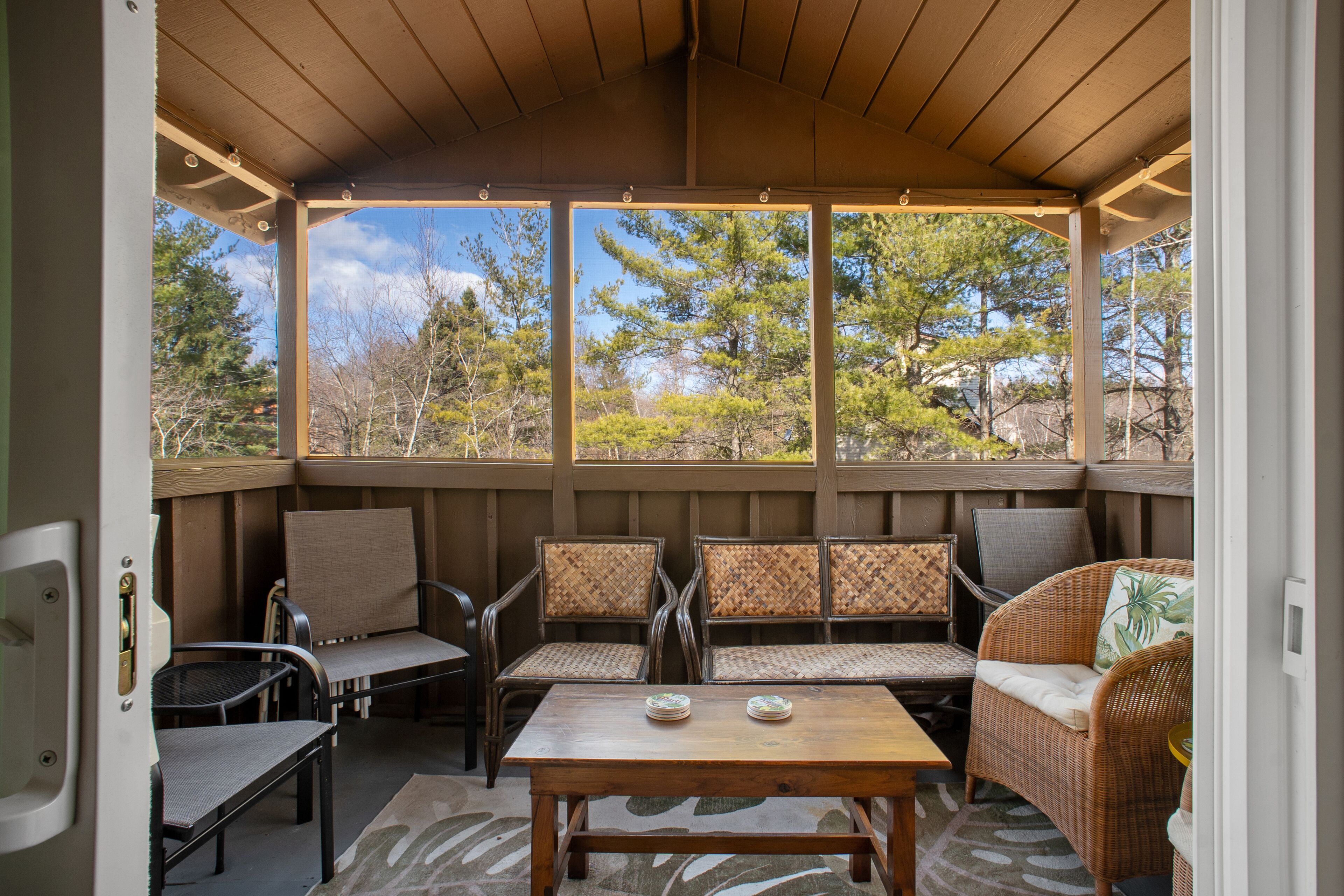 A covered patio with wicker furniture, a wooden table, and a view of trees.