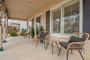 Eine überdachte Terrasse mit Korbstühlen und einem kleinen Tisch, große Fenster mit Jalousien und eine sichtbare amerikanische Flagge.