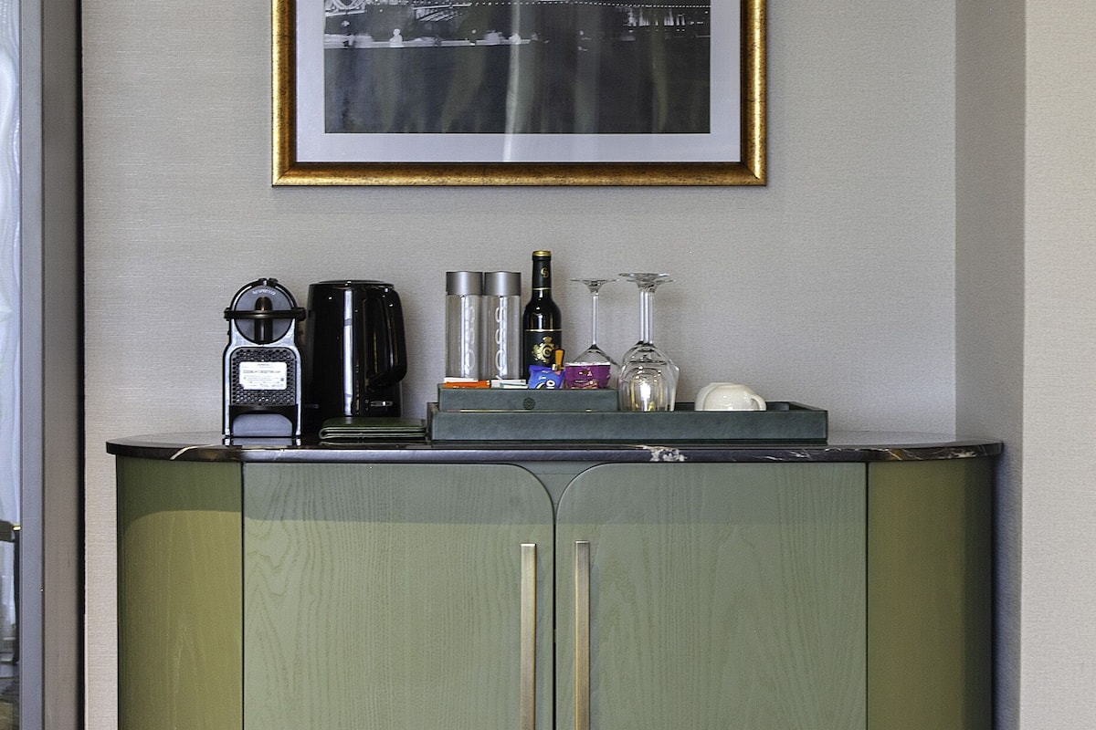 A green cabinet with a framed picture of a bridge, a coffee maker, and wine glasses on top.