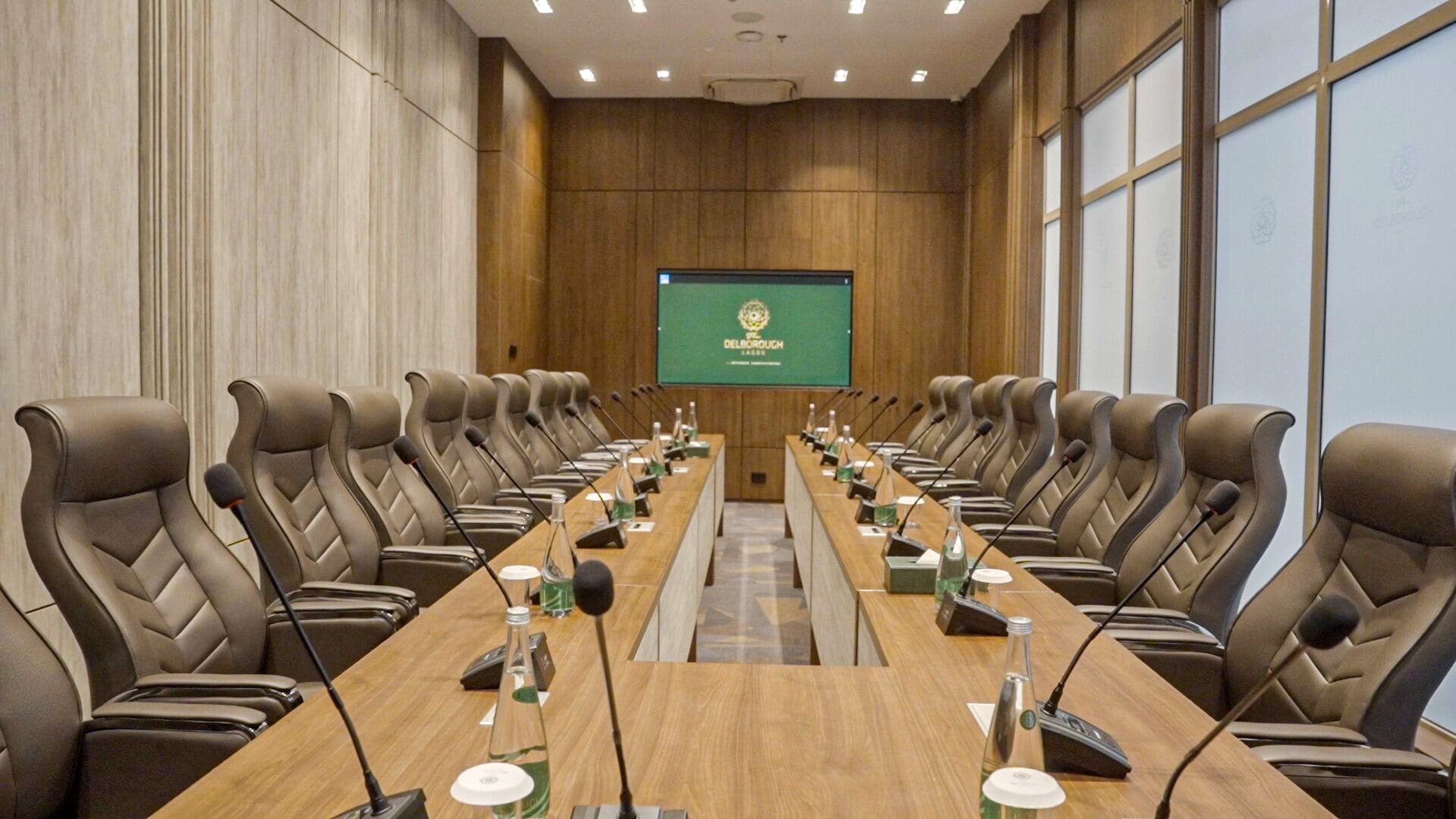 Un salón de conferencias con una larga mesa, numerosas sillas, micrófonos y una pantalla que muestra el logotipo de Delborough.
